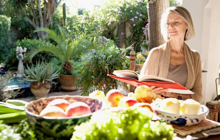 Mulher idosa lendo um livro de receitas com vários alimentos na mesa