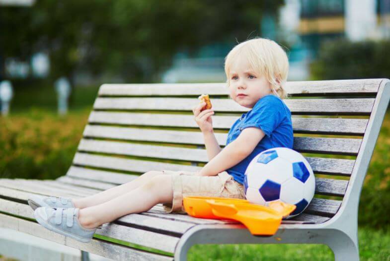 Criança comendo um lanche ao lado de uma bola de futebol