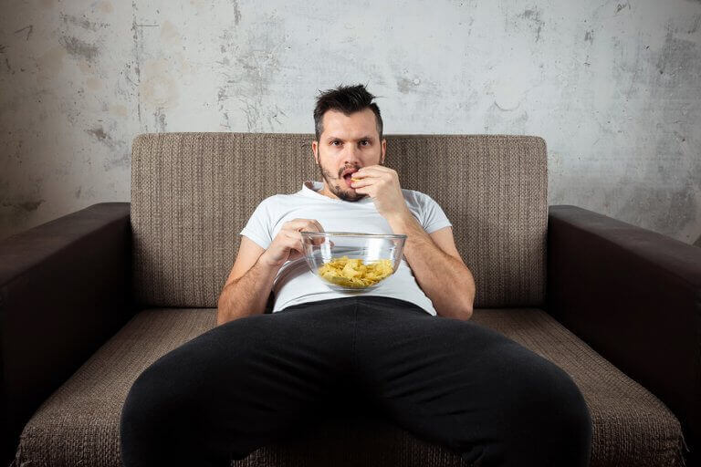 A man eating junk food while sitting on the couch to exemplify a sedentary lifestyle as one of the causes for bad cholesterol