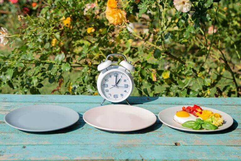 Two empty plates next to a full plate in front of a clock to illustrate intermittent fasting