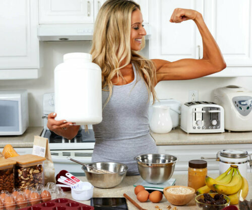 Woman in the kitchen, flexing while holding protein powder with protein rich foods on the counter.