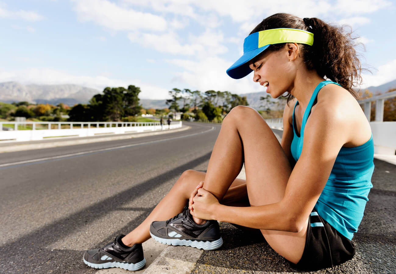 A female marathon runner sitting on the side of the road holding onto her ankle.